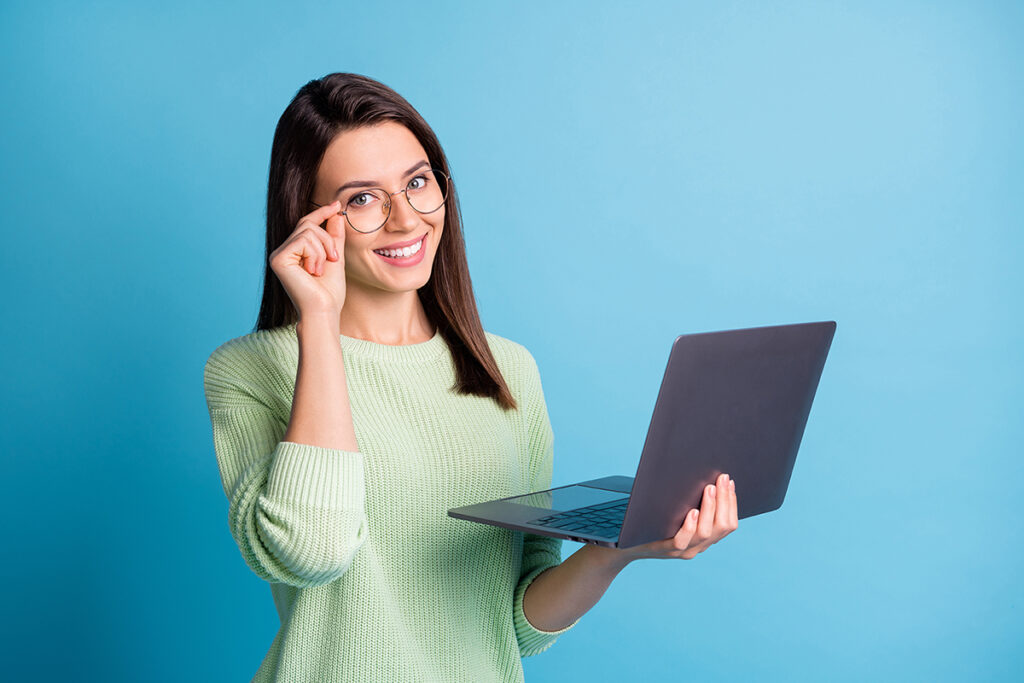 Frau in Pullover hält mit einer Hand den Bügel ihrer runden Brille und mit der anderen Hand einen Laptop