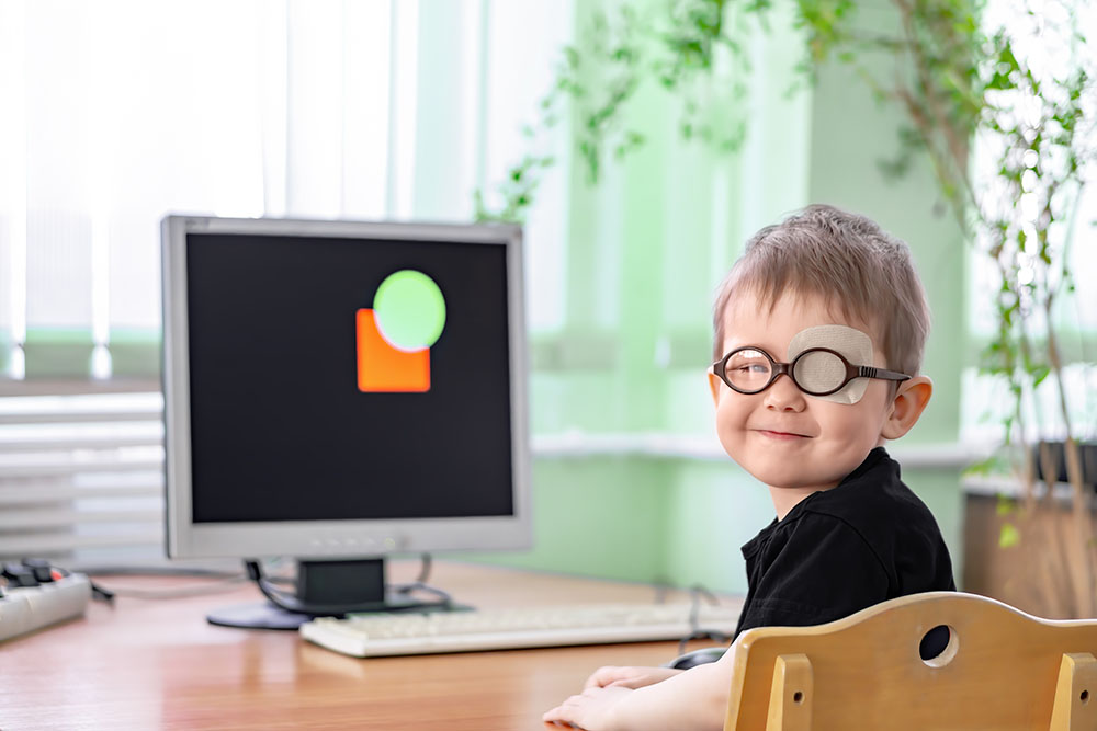 Kleiner Junge mit einer Brillen und einem abgedeckten Auge vor einem Bildschirm sitzend