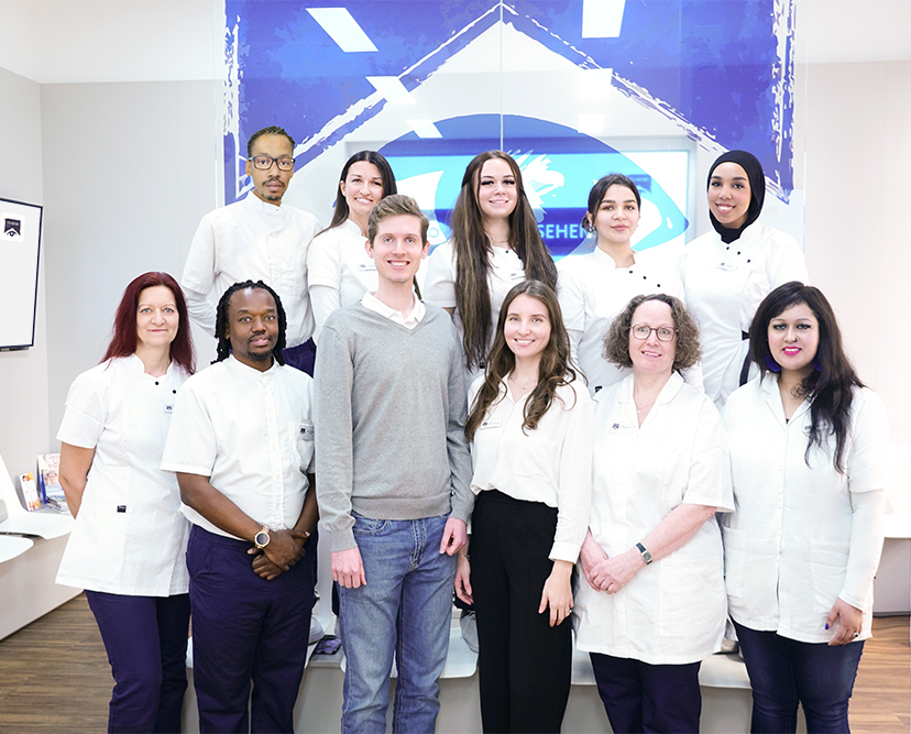 MitarbeiterInnen einer Augenarztpraxis vor Glaswand (mit Logo) und Bilschirm