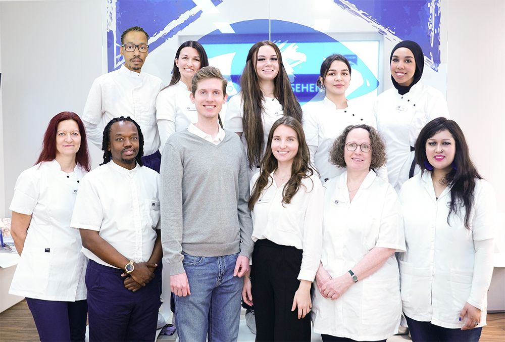 MitarbeiterInnen einer Augenarztpraxis vor Glaswand (mit Logo) und Bilschirm